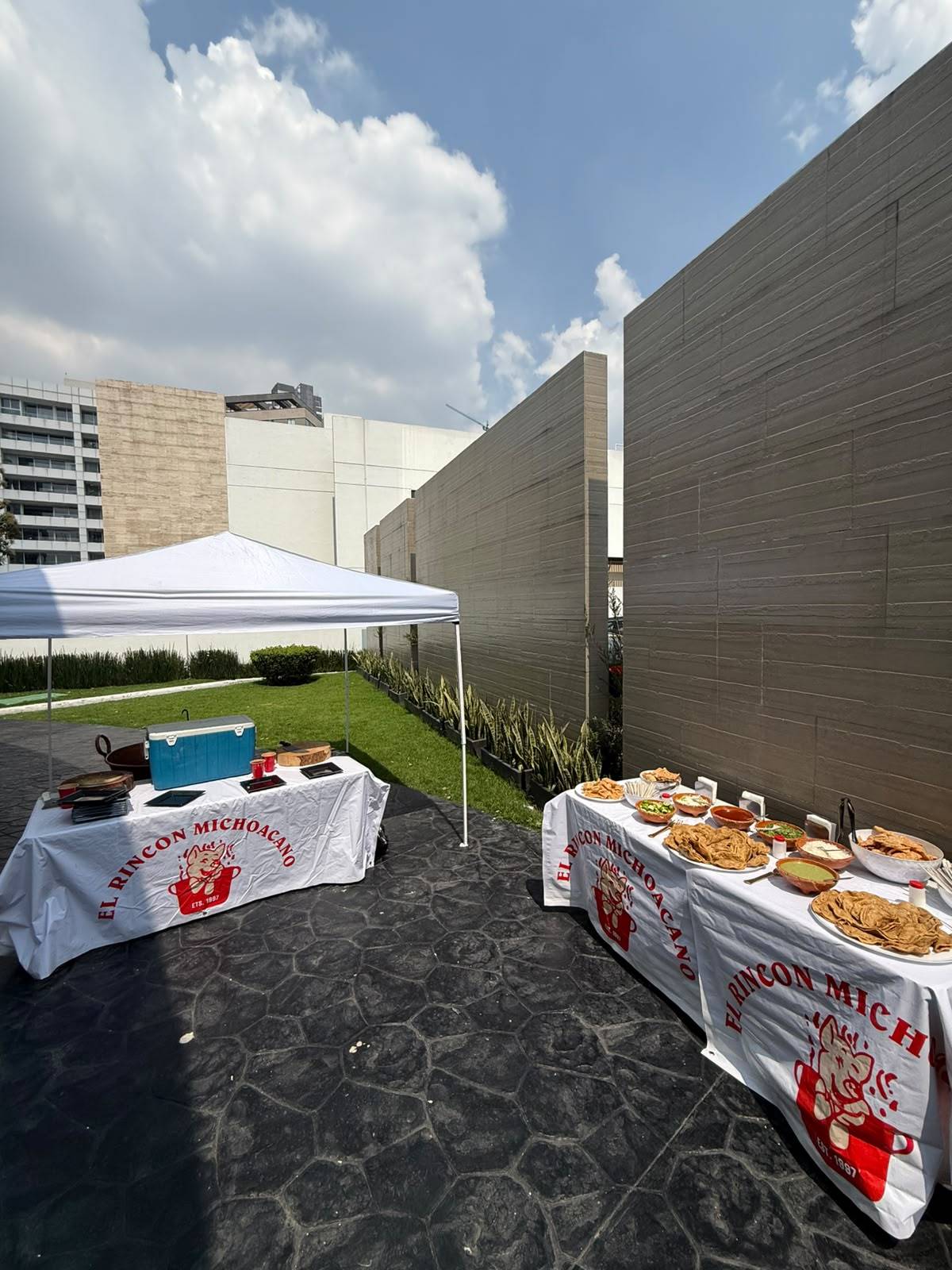 Carpa blanca y dos mesas con manteles del logo, listas para servir carnitas en una terraza